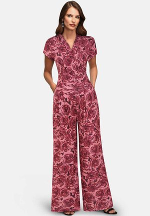 Ženska, oblečena v rožnato širokorezo kombinezo z rdečimi vzorci vrtnic, kratkimi rokavi in rokami v žepih, stoji ob enobarvnem ozadju.