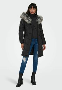 Doudoune noire avec capuche en fausse fourrure, texture matelassée et fermeture éclair. Associée à un jean bleu déchiré et des bottes noires à talons hauts.