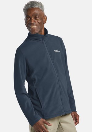 Älterer Mann mit grauen Haaren, der eine dunkelblaue Fleecejacke mit Reißverschluss und beige Hosen trägt, lächelt und schaut zur Seite.