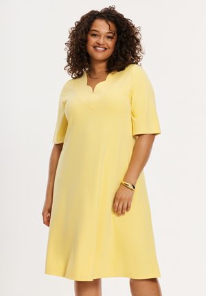 Smilende kvinde med krøllet hår iført en gul kjole til knæet med korte ærmer og en bølget V-hals, accessoriseret med et guldarmbånd.
