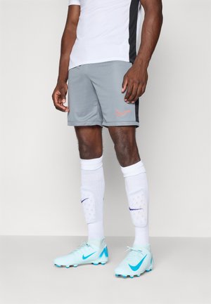 Grå sportsshorts med orange Nike-logo, parret med hvide sokker. Blå og hvide fodboldstøvler har et fremtrædende Nike-swoosh-design.