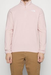 Pullover in pile rosa chiaro con collo a mezza zip, caratterizzato da un logo bianco sul lato sinistro del petto e maniche lunghe. Tessuto morbido e vestibilità casual.