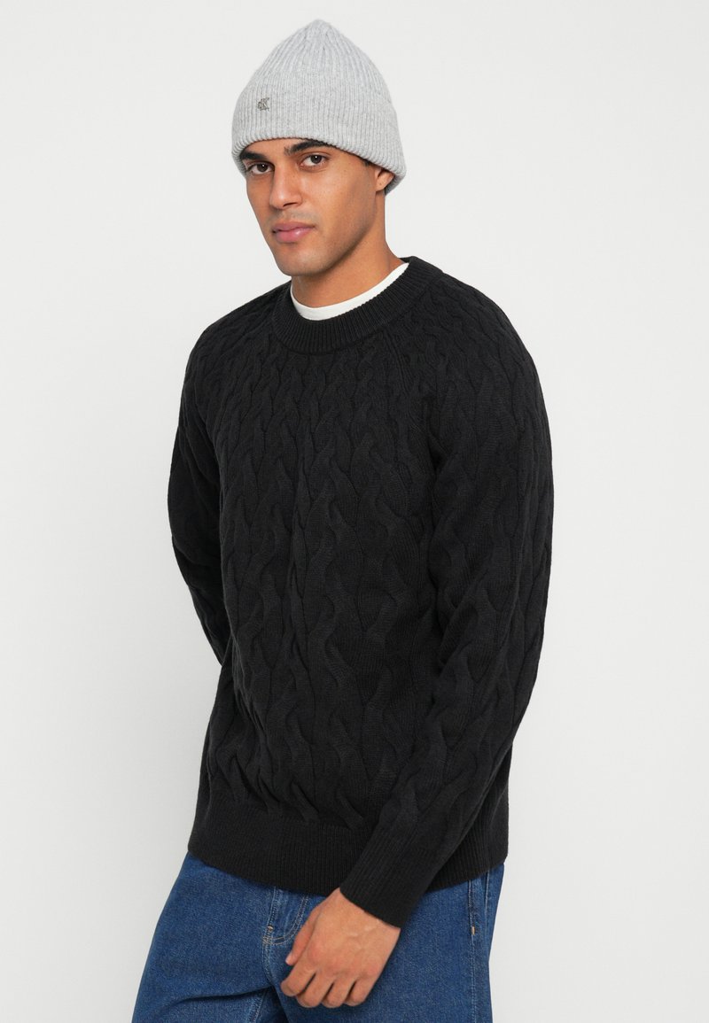 Maglione nero a trecce in materiale spesso; presenta un collo e polsini a coste. Abbinato a un berretto a coste grigio chiaro.