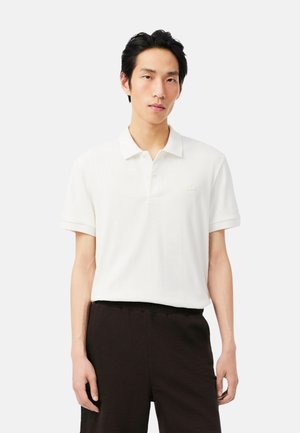 Joven con cabello negro lleva una camisa polo blanca de manga corta y pantalones negros, de pie frente a un fondo claro y sencillo.