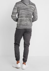 Sudadera gris jaspeada con capucha ajustable, combinada con pantalones cargo oscuros que tienen bolsillos laterales y puños elásticos. Zapatillas blancas visibles.