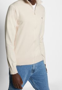 Uomo che indossa un maglione crema con cerniera integrale sopra una camicia bianca abbinata a jeans blu, in piedi contro uno sfondo bianco uniforme.