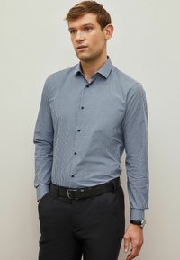 Cămașă cu guler, cu mâneci lungi, în carouri albastre și albe, cu nasturi negri, purtată cu pantaloni întunecate și o curea texturată neagră.