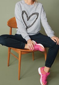 Osoba sedí s nohama zkříženýma na dřevěné židli, má na sobě šedou mikinu s leopardím potiskem ve tvaru srdce a jasně růžové tenisky.