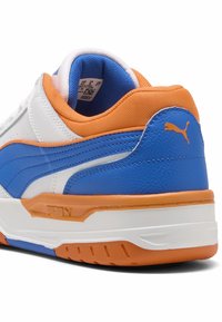 Zapatilla blanca y azul con acentos naranjas, que cuenta con una parte superior de cuero texturizado, un cuello acolchado y una suela de goma con ranuras de tracción.