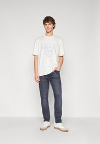 Camiseta blanca de manga corta con texto gráfico en púrpura, combinada con jeans oscuros y zapatillas blancas con suelas y cordones beige.