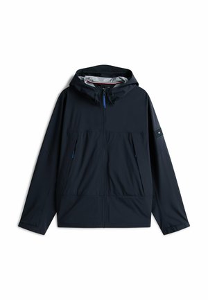 Chaqueta ligera de color azul oscuro con capucha, cremallera frontal, dos bolsillos laterales y puños elásticos. Presenta cremalleras azul contrastante y un parche con el logo.