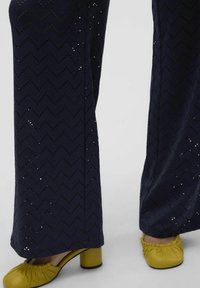 Siniharma kuviolliset leveälahkeiset housut, joissa on sahalaitamainen kuvio, sisältävät aukkoja. Keltaiset korkokengät, joissa on pyöreä kärki ja paksu korko, täydentävät asua.