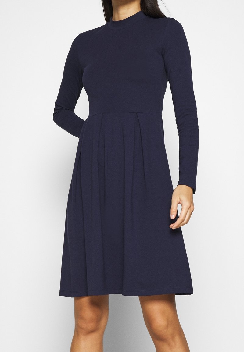 Granatowa sukienka z długim rękawem, okrągłym dekoltem i plisowanym dołem. Wykonana z miękkiego materiału, ma dopasowaną górę i rozszerzający się dół.