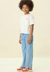 Witte t-shirt met korte mouwen en rode hartdetail, lichtblauwe wijde jeans, en meerkleurige sneakers met textuuraccenten.