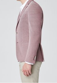 Blazer texture rosso chiaro con chiusura a un bottone, tasca a toppa e dettaglio di spacco laterale, abbinato a una camicia bianca e pantaloni beige.