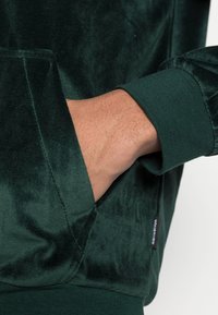 Grön sammets hoodie med en kenguruficka, ribbad mudd och en etikett på sidan. Tyget har en mjuk, texturerad yta.