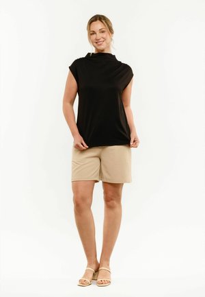 Kobieta stojąca, ubrana w czarny top bez rękawów, beżowe szorty do kolan oraz beżowe sandały, na jednolitym białym tle.