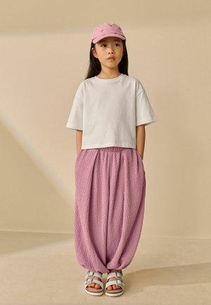 Ung pige står med hænderne i lommerne iført hvid kortærmet skjorte, teksturerede løse lyserøde bukser, lyserød kasket og hvide sandaler.