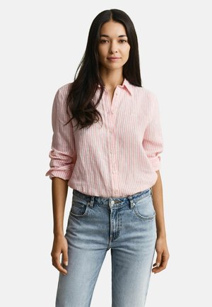 Frau mit langen dunklen Haaren, die ein rosa-weiß gestreiftes, geknöpftes Hemd mit hochgekrempelten Ärmeln und hellblaue Jeans trägt, steht vor einem weißen Hintergrund.