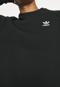 Svart sweatshirt gjord av mjukt tyg. Rund hals, ribbade cuffs, pryds av liten vit Adidas-logotyp på bröstet. Slät textur, inga mönster.