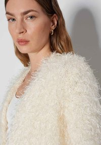 Cardigan in maglia morbida e fluttuante di un bianco sporco, con un aspetto testurizzato e morbido. I lunghi filamenti creano un effetto ciuffo. Presenta un design semplice e aperto.