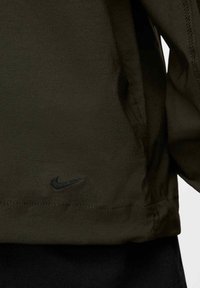 Nike-jacka i mörkgrön tyg, med en svart broderad logotyp, rak passform och strukturerad yta med synliga sömnadsdetaljer.