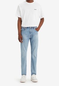 La personne porte un t-shirt blanc Levi’s, un jean en denim bleu clair et des baskets blanches, debout avec une main dans la poche sur fond blanc.