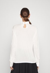 Vit långärmad skjorta med en volangkrage och nyckelhålsutskärning på baksidan, med knäppta manschetter och en mjuk, slät textur.