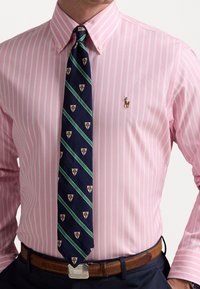 Světle růžová pruhovaná košile s l ímečkem na knoflíky, doplněná navy kravatou se zelenými diagonálními pruhy a vzory insignií. Hnědý opasek.