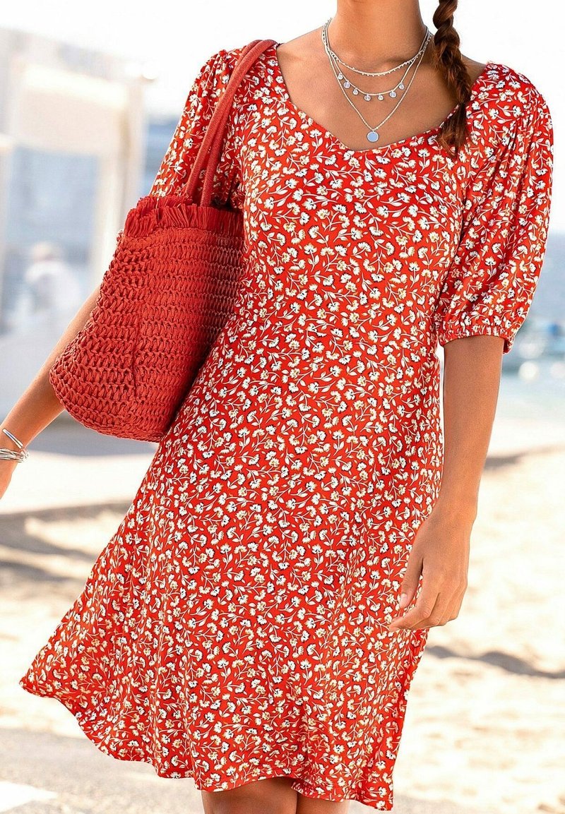 Femme portant une robe rouge à fleurs avec des manches bouffantes, portant un sac fourre-tout rouge texturé et des colliers argentés superposés en plein air.