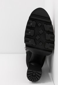 Semelle d'une botte noire avec une épaisse bande de roulement à motifs, inclinée contre un angle entre un mur blanc et un sol, mettant en avant le design texturé de la semelle antidérapante.