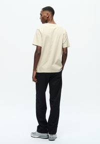 Beige bomulds t-shirt med korte ærmer, rund halsudskæring og afslappet pasform, parret med sorte bukser og grå sportsko. Minimalistisk design.