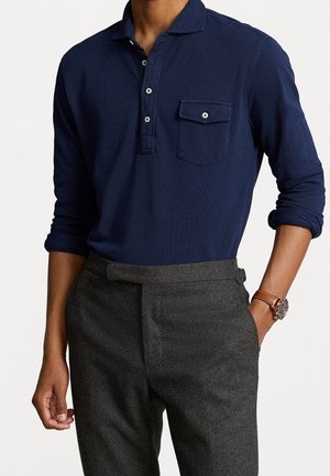 Camisa polo de manga larga en azul marino con cuello, tapeta de tres botones y bolsillo en el pecho, combinada con pantalones grises a medida y un reloj marrón.