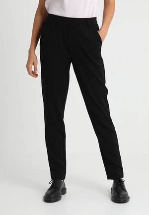 Persoon draagt nauwsluitende zwarte broek met zijzakken en zwarte veterschoenen, staand tegen een effen witte achtergrond.