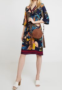 Blommönstrad knälång klänning i marinblått med färgglada tropiska blad. Kombineras med en rund brun mockaväska med fransdetalj.