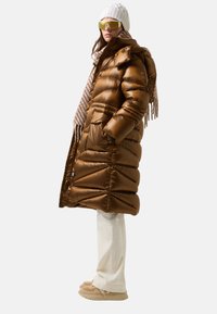 Kobieta w brązowym, pikowanym długim płaszczu, z kremową wełnianą czapką, beżowym paskiem w paski, białych spodniach i beżowych butach wsuwanych, stojąca bokiem.