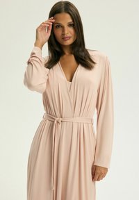 Różowa sukienka kopertowa z długimi rękawami, głębokim dekoltem w kształcie V i paskiem w talii. Materiał jest gładki i zwiewny, z eleganckimi fałdami.