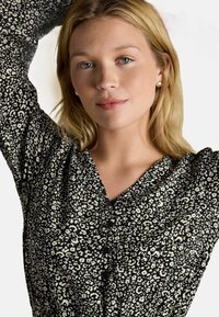 Blonde Frau mit hellblauen Augen trägt Perlenohrringe und eine schwarz-weiße gemusterte Bluse mit Knöpfen und posiert mit erhobenen Armen.