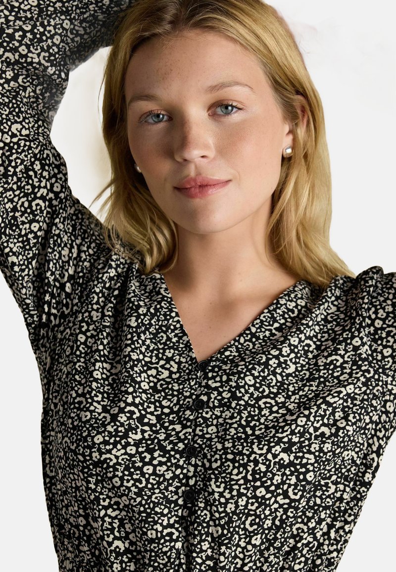 Blonde Frau mit hellblauen Augen trägt Perlenohrringe und eine schwarz-weiße gemusterte Bluse mit Knöpfen und posiert mit erhobenen Armen.