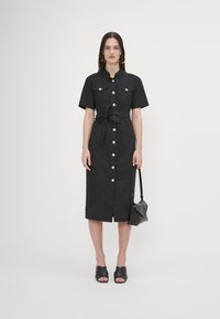 Rochie neagră cu mâneci scurte, cu nasturi, talie marcata cu un brâu, două buzunare pe piept, lungime până la genunchi, și nasturi aurii. Asortată cu o geantă neagră și pantofi cu vârful deschis.