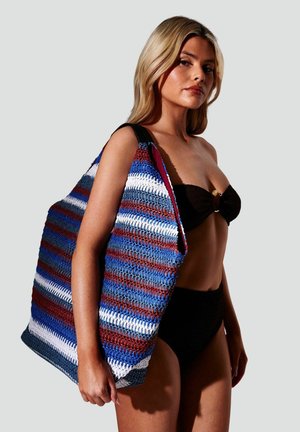 Donna in bikini nero che porta una grande borsa a tracolla in crochet con strisce blu, bianche e marroni su spalla contro sfondo grigio chiaro.
