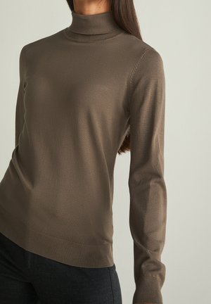 Mujer con un suéter entallado de cuello alto color topo, de manga larga con puños y bajo de canalé, sobre un fondo claro y liso.