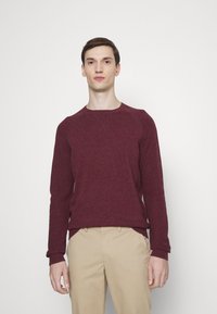 Bordeauxfarbenes Sweatshirt mit Rundhalsausschnitt, gerippten Bündchen und Saum, strukturiertem Strickstoff. Getragen mit beigen Hosen, minimalistisches Design, neutraler Hintergrund.