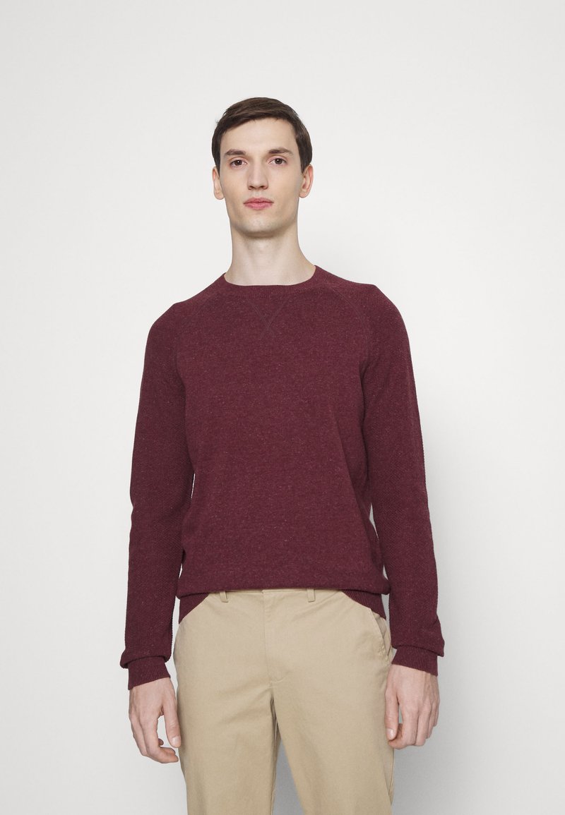 Bordeauxfarbenes Sweatshirt mit Rundhalsausschnitt, gerippten Bündchen und Saum, strukturiertem Strickstoff. Getragen mit beigen Hosen, minimalistisches Design, neutraler Hintergrund.