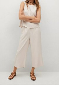 Femme portant un top beige sans manches et un pantalon beige à jambes larges, associé à des sandales plates brunes à lanières, se tenant les bras croisés.