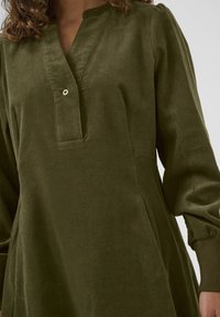 Olijfgroen corduroy jurk met een V-hals en knoopdetail. Heeft pofmouwen en zijzakken. De textuur is zacht en ribbelig.