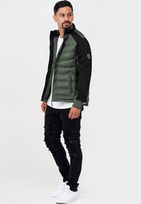 Chaqueta inflable verde con mangas negras, cierre de cremallera y puños acanalados. Llevada con jeans negros desgastados y zapatillas blancas.