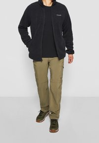 Giacca in pile nera, pantaloni cargo verde oliva con zip e scarpe da trekking verde scuro con dettagli arancioni. Texture liscia e vestibilità casual.