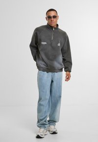 Pullover in felpa grigia con zip a metà e finitura testurizzata, caratterizzato da dettagli del logo a contrasto. Abbinato a jeans chiari e sneakers.