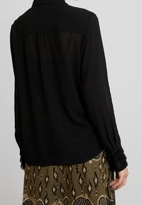 Svart genomskinlig blus med långa ärmar, med en rynkad yoke och texturerade fransar på ärmsluten, i kombination med en mönstrad olivgrön kjol.